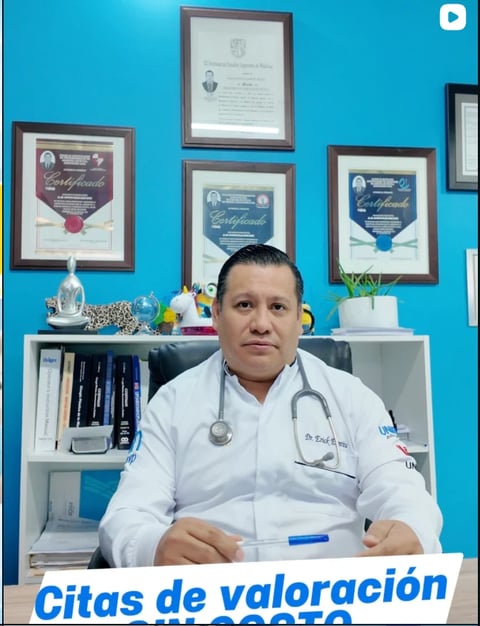 Doctor in white coat with stethoscope sitting at desk in front of blue wall with framed certificates