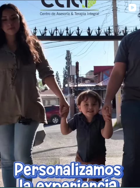 A young child holds hands with two adults on a sunny street, with a therapy center sign visible in the background