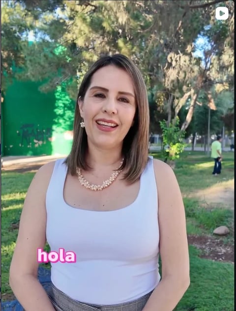 Woman smiling at camera in white sleeveless top with pearl necklace, standing in park with trees and grass behind her
